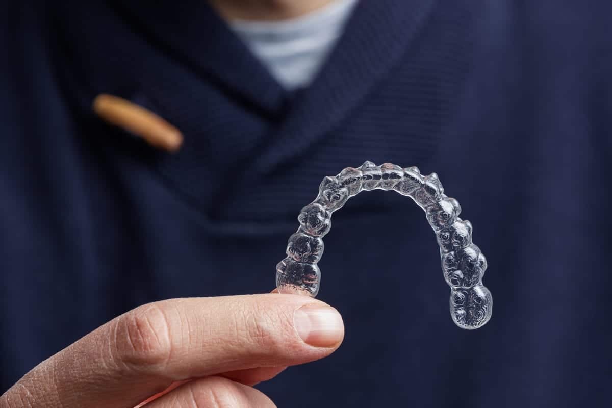 Close-up of a clear Invisalign aligner held on a finger against dark clothing.